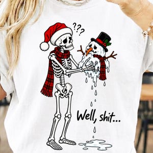 May include: A white t-shirt featuring a graphic of a skeleton wearing a red Santa hat and a red plaid scarf, holding a melting snowman with a black top hat and a red plaid scarf. The text "Well, shit..." is printed below the melting snowman.