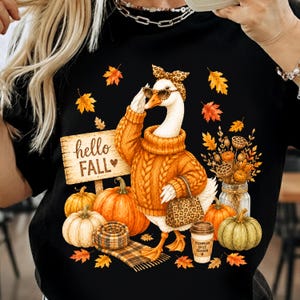 May include: A black t-shirt featuring a graphic of a white goose wearing an orange cable-knit sweater, leopard print headband, and sunglasses. The design includes pumpkins, a wooden sign reading hello FALL, and a cup labeled PUMPKIN SPICE SEASON.