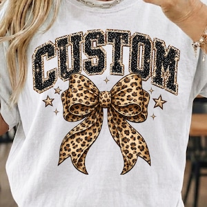 May include: White t-shirt featuring the word "CUSTOM" in a black glitter font with a leopard print border. A large leopard print bow is centered below the word, with star and diamond accents.