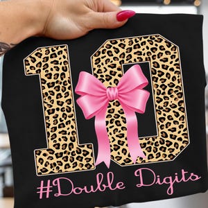 May include: Black t-shirt with a leopard print "10" design and a pink bow. The text "#Double Digits" is in pink script below the numbers. The shirt is held up by a hand.