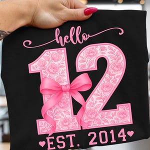 May include: Black t-shirt with the text "hello 12 EST. 2014" in pink. The number 12 is filled with a floral pattern and a pink bow. The shirt is held by a hand.
