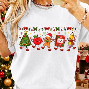以下が含まれることがあります： 白いTシャツに、装飾されたクリスマスツリー、赤いリンゴ、サンタ帽をかぶったジンジャーブレッドマン、赤いノート、黄色い鉛筆という5つのアニメ風クリスマスキャラクターが、クリスマスライトの下に描かれたカラフルなグラフィックがプリントされています。
