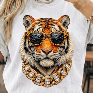 Könnte beinhalten: Weißes T-Shirt mit einer detaillierten Illustration eines Tigers, der eine Sonnenbrille mit Leopardenmuster und eine goldene Kette mit großen quadratischen Edelsteinen trägt. Das Fell des Tigers ist orange und schwarz. Das Design ist auf dem T-Shirt zentriert.