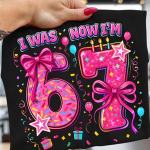 以下が含まれることがあります： 「I WAS NOW I'M 67」の文字が入った黒いTシャツ。数字の6はスプリンクル付きドーナツのようにデザインされ、数字の7はグリッターで装飾されています。ピンクのリボン、星、風船、紙吹雪がデザインを完成させています。