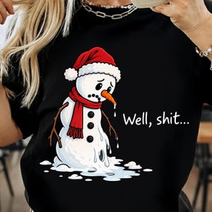 May include: A black t-shirt featuring a graphic of a melting snowman wearing a red hat and scarf, with a sad expression and an orange carrot nose. The text "Well, shit..." is printed in white next to the snowman, which is pooling at the base.