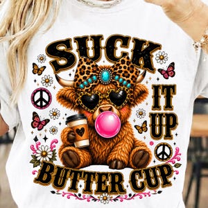 May include: A white t-shirt featuring a graphic of a brown highland cow wearing a leopard print bow with a turquoise stone, heart-shaped sunglasses, and blowing a pink bubble. The cow holds a coffee cup. Text reads SUCK IT UP BUTTER CUP.