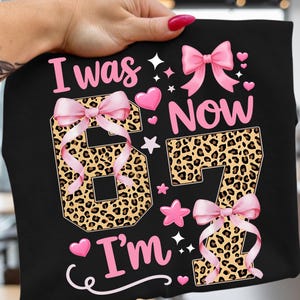 以下が含まれることがあります： 黒いTシャツにピンク色の「I was 67 Now I'm 67」の文字。数字はヒョウ柄で、ピンクのリボンが付いています。ピンクのハートと星がシャツを飾っています。