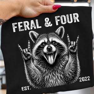 Pode incluir: Uma t-shirt preta com uma ilustração gráfica branca de um guaxinim com a boca aberta e a língua de fora, fazendo um gesto de mão de rock and roll com ambas as patas. O texto "FERAL & FOUR" está impresso acima, com "EST. 2022" abaixo.