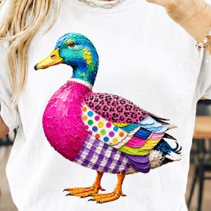 May include: A white t-shirt featuring a colorful mallard duck graphic with a textured, impasto-style paint effect. The duck has a blue head, a bright yellow beak, a vibrant pink chest, a leopard print wing, polka dot accents, and a purple gingham pattern.