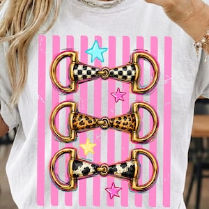 Puede incluir: Camiseta blanca con tres diseños de bocados de caballo con detalles dorados y centros estampados. Los estampados incluyen un tablero de ajedrez en blanco y negro, estampado de leopardo y diseño de piel de vaca. Rayas verticales rosas y estrellas completan el diseño.