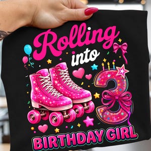 Może przedstawiać: Czarny t-shirt z napisem "Rolling into Birthday Girl". Koszulka przedstawia różowe rolki, cyfrę 3 ze świeczkami, sercami, gwiazdkami i kokardą. Projekt jest w kolorze różowym, białym i niebieskim.