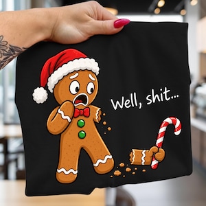May include: A black t-shirt featuring a graphic of a brown gingerbread person wearing a red and white Santa hat and a red bow tie. The gingerbread person has a shocked expression, a missing arm, and is holding a red and white candy cane. The text reads Well, shit...