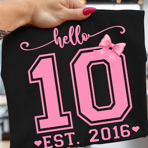 May include: Black t-shirt with the text "hello 10 EST. 2016" in pink. The number 10 is in a bold, outlined font, and a pink bow is on the right side. The shirt is held up by a hand.