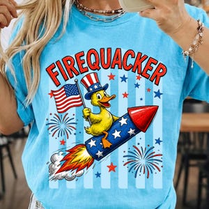 May include: Light blue t-shirt with a patriotic design. The shirt features the word "FIREQUACKER" in red, a yellow duck wearing a patriotic hat, and an American flag. The duck is riding a rocket with stars and fireworks.