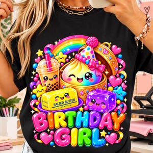 Può includere: Una t-shirt nera con una grafica colorata che raffigura un arcobaleno, un tè boba, un panetto di burro sorridente con l'etichetta Salted Butter 113 g, un cubo viola e un cappellino da festa. Il testo Birthday Girl è stampato in grandi lettere.
