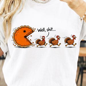 May include: A white t-shirt featuring a graphic of an orange pie shaped like a video game character chasing three brown turkeys. The first turkey has a speech bubble above its head with the black text "Well, shit..." printed in a simple font.
