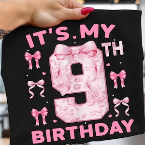 以下が含まれることがあります： ピンク色の文字で「IT'S. MY 9TH BIRTHDAY」と書かれた黒いTシャツ。数字の9は花柄とピンクのリボンで装飾されています。追加のピンクのリボンとハートが数字の周りに散らばっています。
