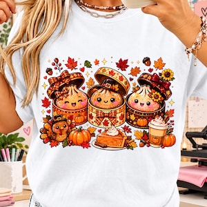 Könnte beinhalten: Ein weißes T-Shirt mit einer bunten Grafik von drei lächelnden Teigtaschen in gemusterten Geschenkboxen, einem kleinen Truthahn mit Pilgerhut, einem Stück Kürbiskuchen mit Sahne, einem Kaffeebecher, Kürbissen und Herbstblättern.