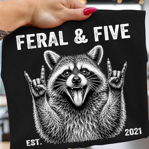 Può includere: Una t-shirt nera con un'illustrazione grafica bianca di un procione con la bocca aperta e la lingua di fuori, che fa un gesto rock and roll con entrambe le zampe. Il testo FERAL & FIVE è stampato sopra e EST. 2021 è stampato sotto.