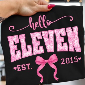 May include: Black t-shirt with the words "hello ELEVEN" in pink script and block letters. The word "ELEVEN" is filled with a floral pattern. A pink bow and the year "2015" are also featured.