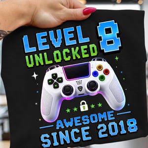 May include: Black t-shirt featuring a white video game controller. The text says "LEVEL 8 UNLOCKED AWESOME SINCE 2018". The controller has coloured buttons and a purple and blue gradient.