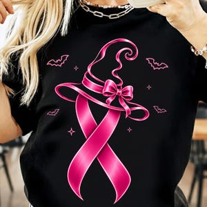 以下が含まれることがあります： 黒いTシャツで、リボンと蝶結びで魔女の帽子を形作った鮮やかなピンク色の啓発リボンのグラフィックが描かれています。デザインには、中央のリボンと帽子のイラストの周囲に小さなピンクのコウモリと四角い星のアクセントが散りばめられています。