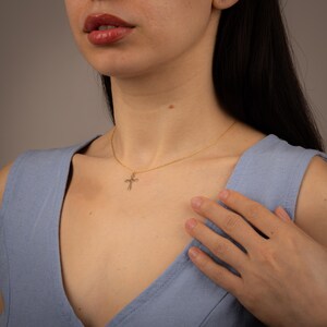 Op de afbeelding: Een gouden ketting met een klein, delicaat kruisje als hanger. De ketting wordt gedragen door iemand met lang, donker haar en een blauwe top zonder mouwen.