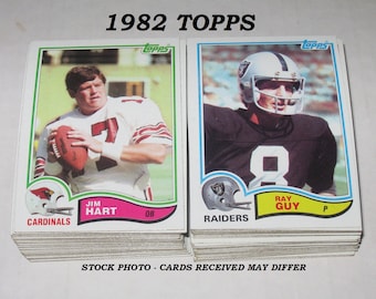 May include: A stack of 1982 Topps football trading cards featuring Jim Hart of the Cardinals and Ray Guy of the Raiders. The cards are in their original packaging.