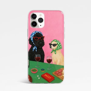 May include: A phone case featuring an illustration of two dogs wearing sunglasses and headscarves, sitting at a table with wine glasses and snacks. The word "SASSY" is on a red box. The background is pink.
