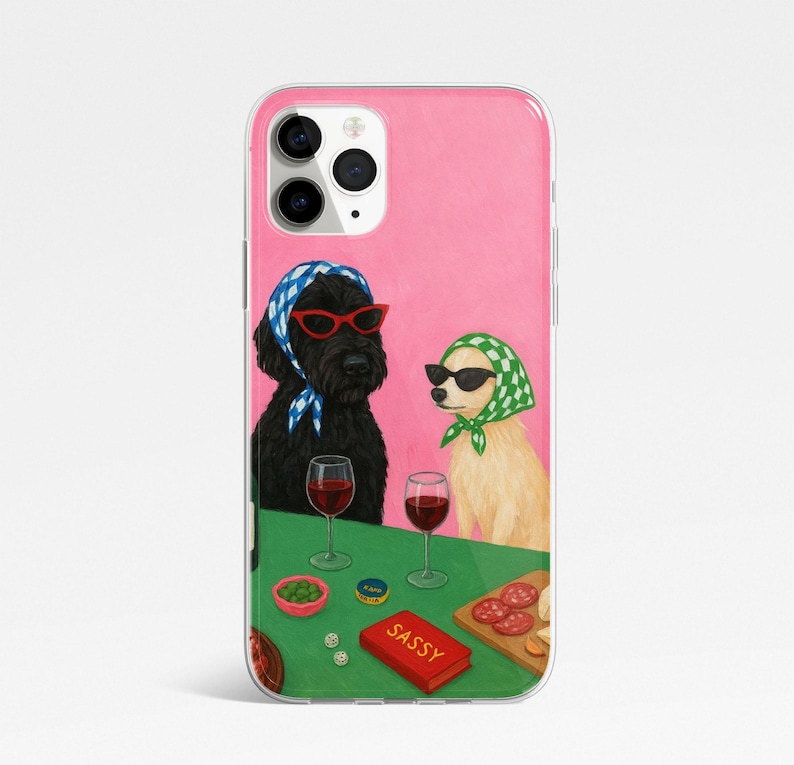 May include: A phone case featuring an illustration of two dogs wearing sunglasses and headscarves, sitting at a table with wine glasses and snacks. The word "SASSY" is on a red box. The background is pink.
