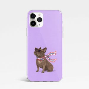 May include: A phone case with a watercolor illustration of a brown French bulldog wearing sunglasses and a red, white, and blue scarf. The case has a lavender background and a clear, glossy finish.
