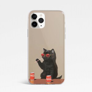 Pu&ograve; includere: Una custodia per telefono trasparente con un gatto nero che indossa occhiali da sole rossi e tiene una cannuccia. Il gatto &egrave; seduto su un tavolo con due lattine di Coca-Cola rosse e bianche.