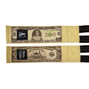 Puede incluir: Dos correas de halterofilia beige con un dise&ntilde;o que se asemeja a un billete de 300 $. Las correas presentan detalles en negro y el texto "THE PERFECT ASS" y "THREE HUNDRED".