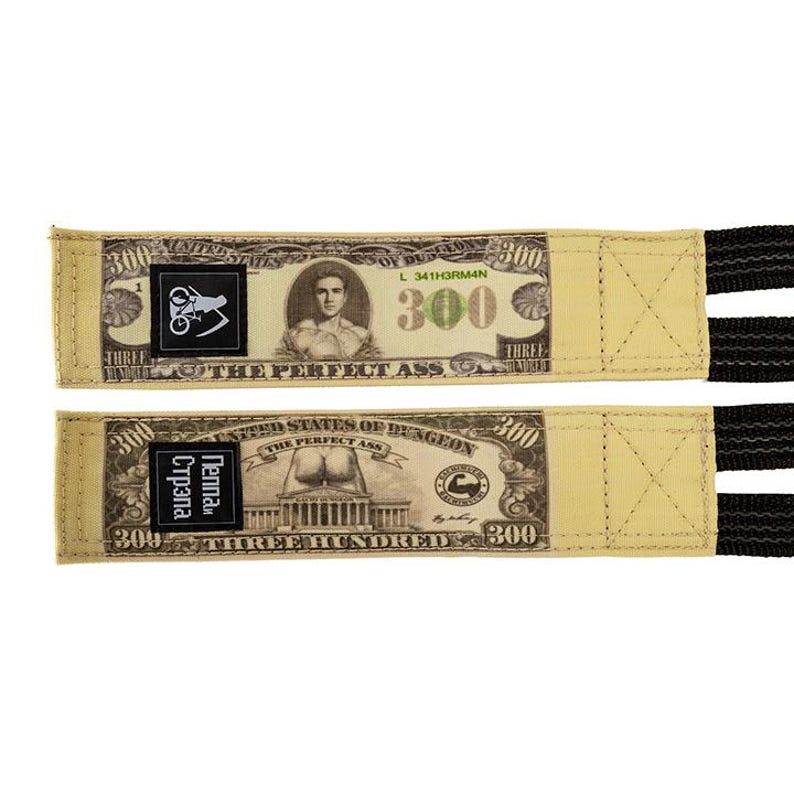 Puede incluir: Dos correas de halterofilia beige con un dise&ntilde;o que se asemeja a un billete de 300 $. Las correas presentan detalles en negro y el texto "THE PERFECT ASS" y "THREE HUNDRED".