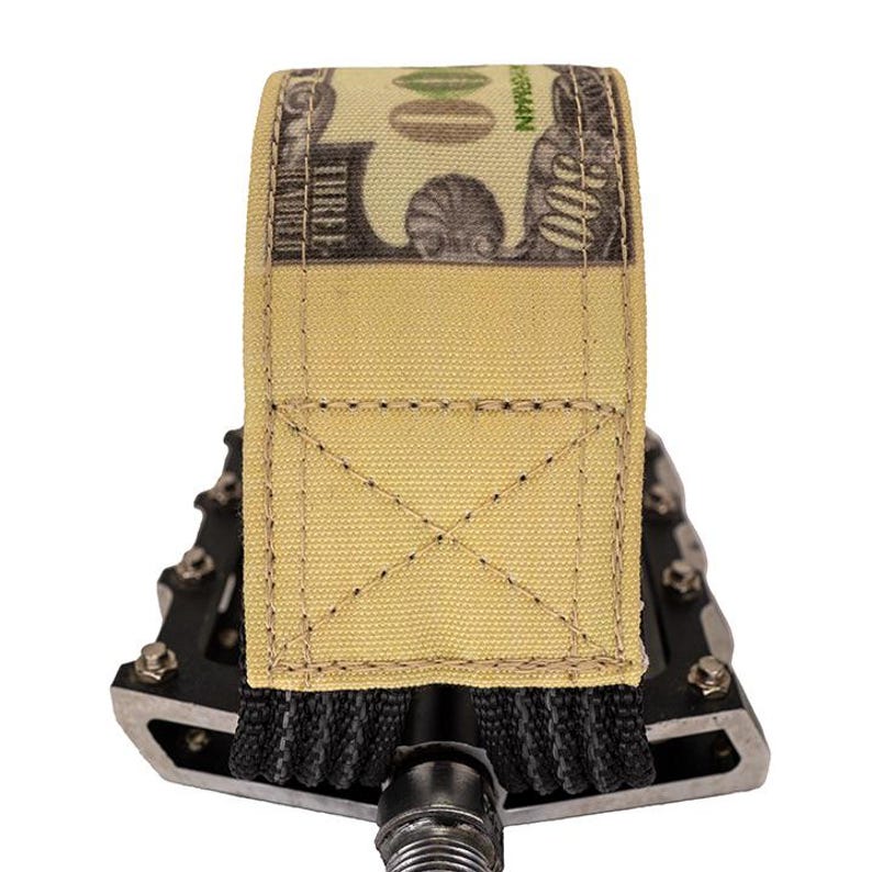 Puede incluir: Una correa de tela color beige con un dise&ntilde;o de billete de 100 $, cosida con un patr&oacute;n de cruz, est&aacute; envuelta alrededor de un pedal de bicicleta negro. El pedal tiene un marco de metal plateado y un agarre negro.