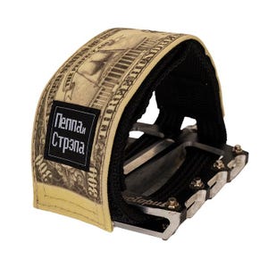 Puede incluir: Un pedal de bicicleta con una correa beige con un dise&ntilde;o de billete de d&oacute;lar. La correa tiene un interior negro y una etiqueta negra con texto blanco. El pedal es plateado con detalles en negro.