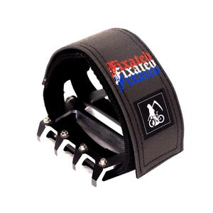 May include: A grey bicycle pedal strap with a black and white logo that says "Fixed" in red, white and blue lettering. The strap is attached to a silver pedal with black accents.