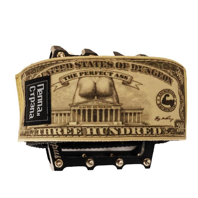 Puede incluir: Un pedal de bicicleta con una envoltura de tela beige que presenta una parodia de un billete de d&oacute;lar. El dise&ntilde;o incluye el texto "THE PERFECT ASS" y "THREE HUNDRED". Una etiqueta negra dice "Pennan Cтрана".