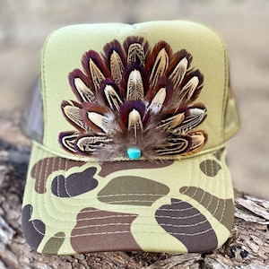 May include: A green camouflage trucker hat with a brown and white pheasant feather accent and a turquoise stone in the center.