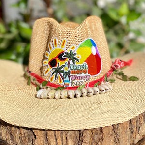 May include: A straw cowboy hat with a red ribbon, green leaves, and white shells around the base. The hat has a patch with a sun, palm trees, a surfboard, and the text "Beach more worry less".