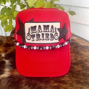 May include: A red trucker hat with a mesh back and a curved brim. The front features a patch with the words "MAMA TRIED" and guitar illustrations. Black star-shaped accents and a beaded band with dice and 8-ball beads add detail.