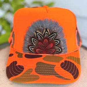 May include: Bright orange trucker hat with a camouflage brim. The hat features a decorative arrangement of feathers in shades of gray, brown, and red. The hat is designed for outdoor activities and adds a touch of style.