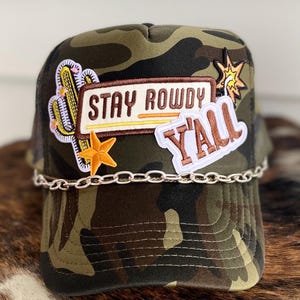 May include: Camouflage trucker hat with a chain detail. The hat has several patches, including one that says "Stay Rowdy Y'all".