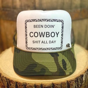 May include: White trucker hat with a camouflage brim. The front of the hat has a black and white rectangular patch with the text "Been Doin' Cowboy Shit All Day."