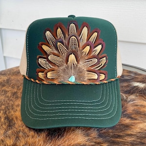 May include: A green trucker hat with a brown and beige pheasant feather design and a turquoise stone accent.
