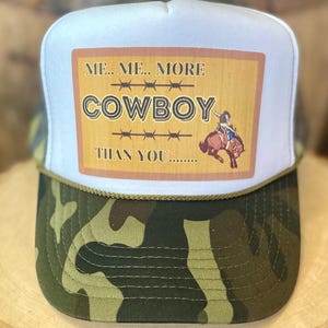 May include: A white trucker hat with a camouflage brim. The front of the hat has a brown and white rectangular patch with the text "ME.. ME.. MORE COWBOY THAN YOU....." and a cartoon image of a cowboy riding a bull.