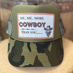 May include: Green camouflage trucker hat with a brown patch that says "Me.. Me.. More Cowboy Than You...." with a skull wearing a cowboy hat and bandana.