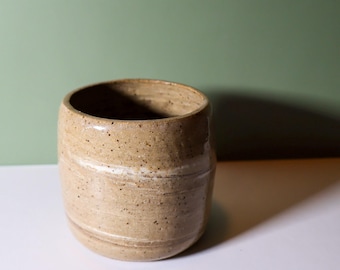 Może przedstawiać: Ręcznie wykonany ceramiczny kubek w odcieniach brązu i beżu. Cylindryczny kubek ma szeroki otwór i zaokrągloną podstawę. Biały pas otacza środek kubka. Tło jest stonowane zielone.
