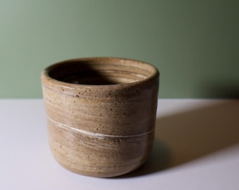 Może przedstawiać: Ręcznie wykonany, cylindryczny ceramiczny kubek z nakrapianym, jasnobrązowym zewnętrzem. Cienki, biały pasek otacza kubek. Kubek ma gładkie, nieszkliwione wykończenie i lekko rozszerzony brzeg. Tło jest jednolite, stonowane zielone.
