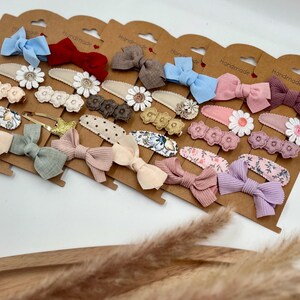 Puede incluir: Un conjunto de 18 pinzas para el pelo hechas a mano con una variedad de colores y patrones. Las pinzas incluyen lazos, flores y lunares. Las pinzas se muestran en una tarjeta marr&oacute;n con la palabra "Handmade" impresa en cada tarjeta.